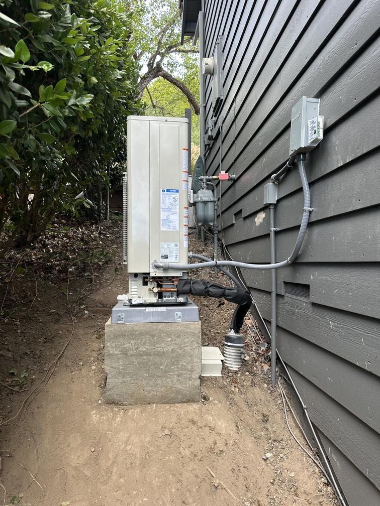 Side-discharge heat pump outdoor unit on a raised pad beside a house, with service disconnect and covered refrigerant lines